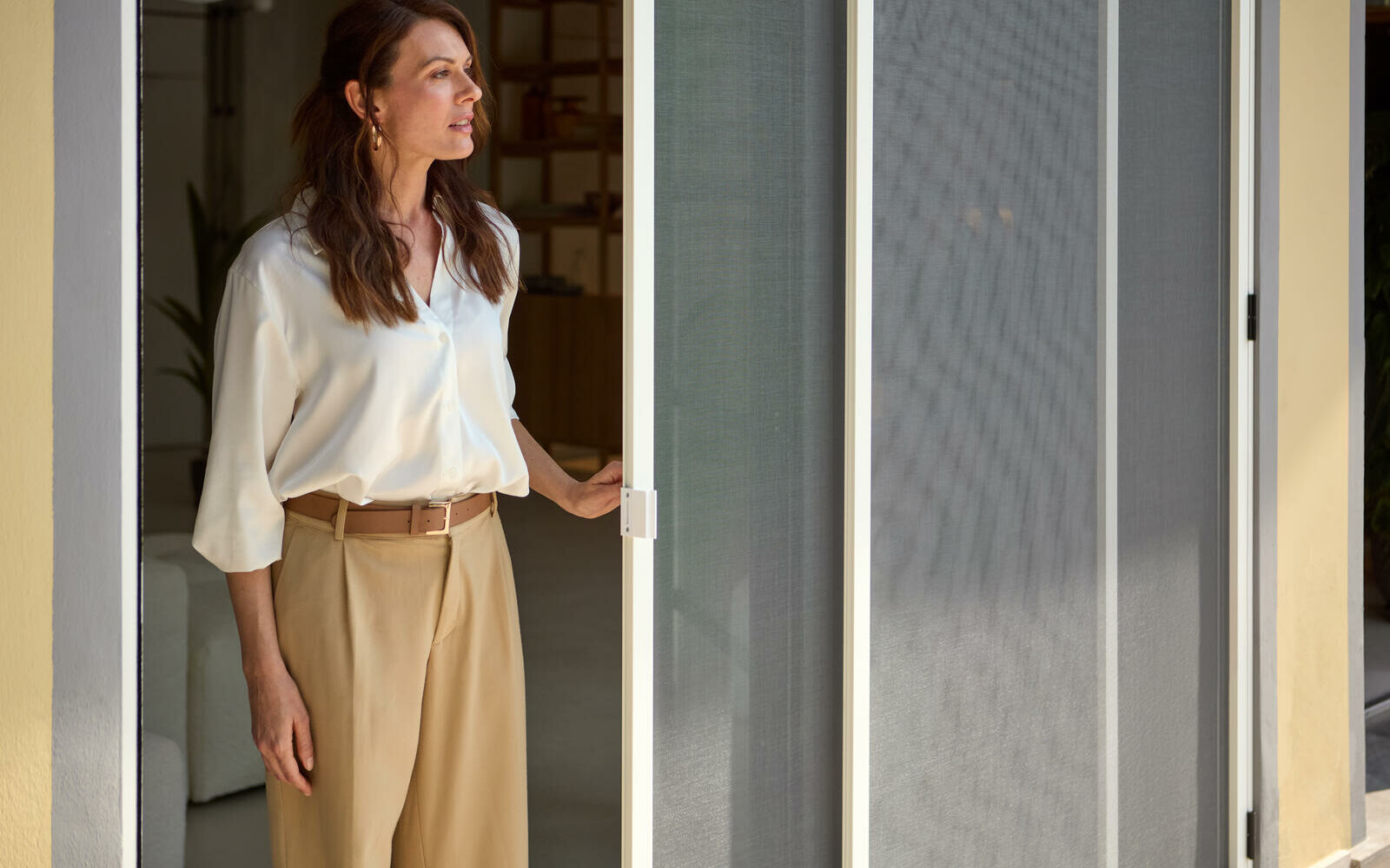 Donna elegante vestita con camicia bianca e pantalone beige in piedi apre zanzariera scorrevole di colore bianco installata in soggiorno mentre guarda verso il giardino esterno della casa