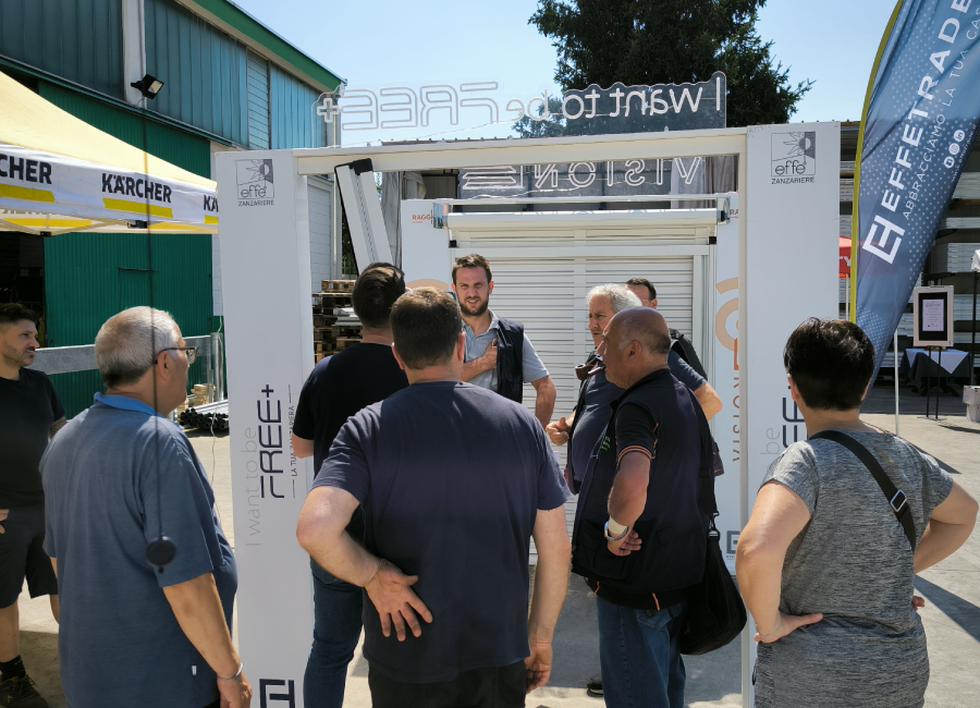 Persone in piedi attorno ad espositori di zanzariere e tapparelle ascoltano le informazioni date da un referente aziendale nel corso di un evento